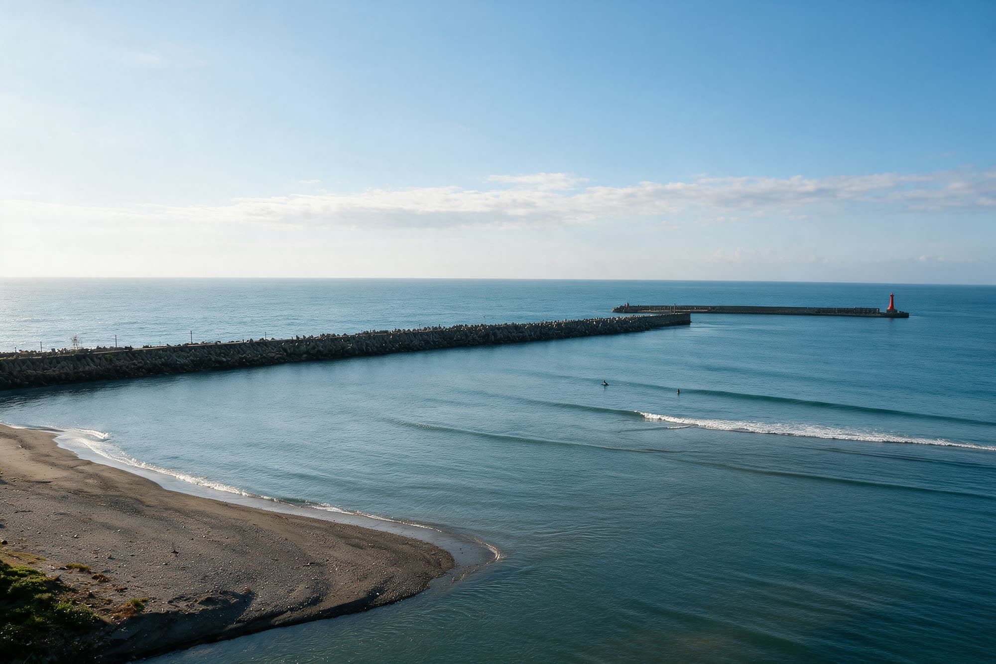 北濱公園外的海灣,長長的消波塊堤防擋在外側,海面平靜,左邊是礫石沙灘,遠處防波堤的盡頭有一座紅色燈塔